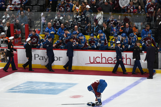 Ein Hockey-Spieler auf dem Eis mit einem Schläger in der Hand, umgeben von einer Gruppe von Menschen mit Mützen und Hockey-Schlägern, mit Zuschauern und Gittern, auf denen "Colorado Avalanche vs San Jose Sharks - 3/24/15 NHL Pick, Odds, and Prediction" steht.
