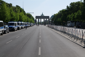 Eine Reihe von Polizeiwagen auf einer Straße vor dem Brandenburger Tor in Berlin, Deutschland geparkt, mit Menschen auf Fahrrädern und auf der Straße stehend, Barrieren, Bäumen und einem Bogen mit Statuen im Hintergrund.