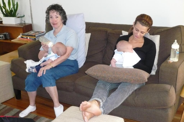 Zwei Frauen sitzen auf einem Sofa und halten Babys, mit Kissen drumherum, in einem Raum mit einer weißen Wand, einem Schreibtisch, Büchern und einer Zimmerpflanze links daneben.