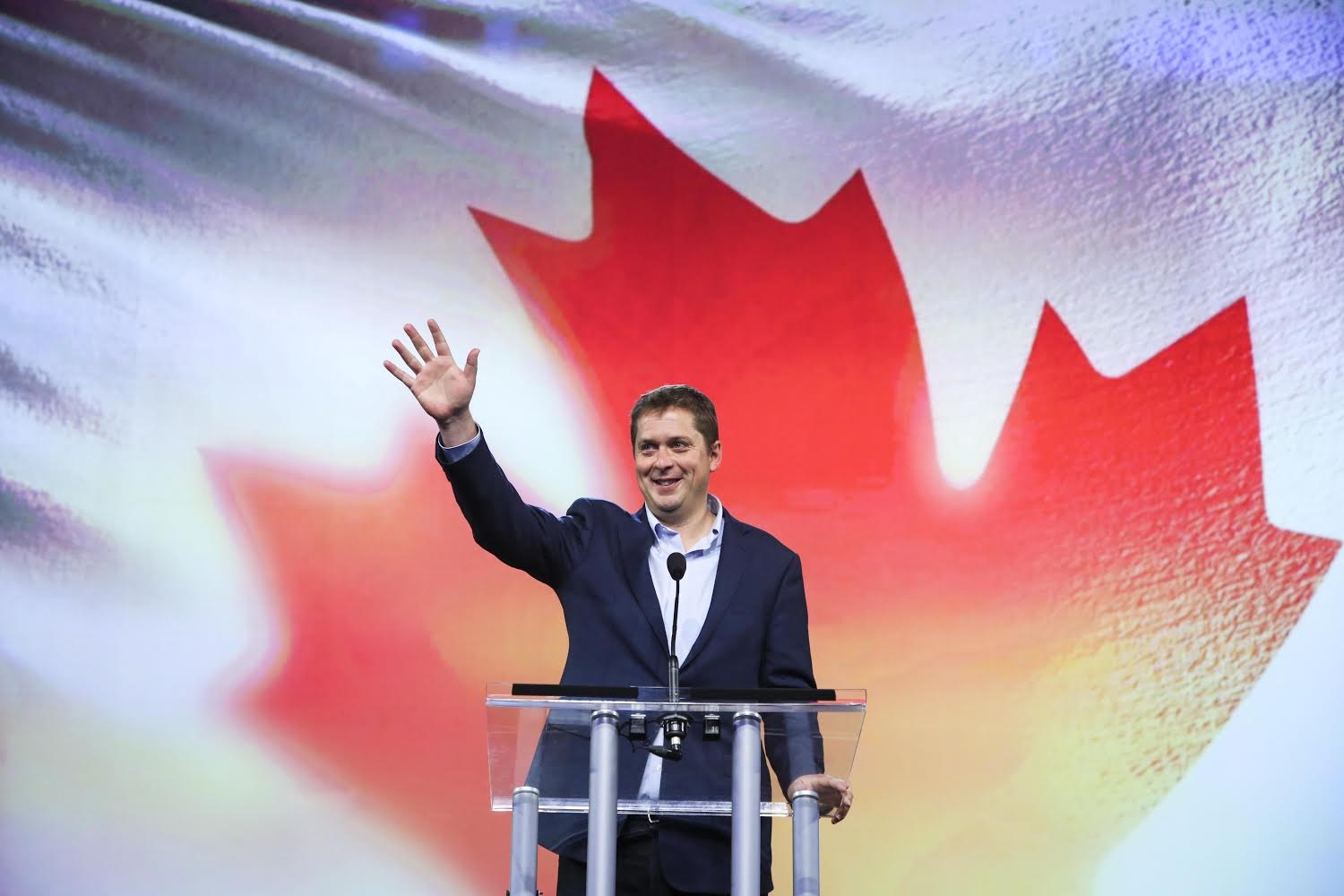 Der kanadische Premierminister Alex Salmond steht an einem Rednerpult mit Mikrofonen, lächelt vor einer kanadischen Flagge, mit einem Bildschirm im Hintergrund.