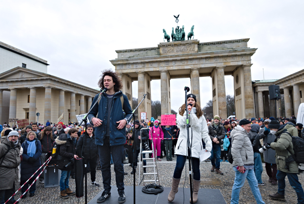 Eine Gruppe von Menschen auf einer Bühne vor dem Brandenburger Tor in Berlin, mit zwei Personen mit Mikrofonen und einer weiteren mit einer Kamera, einer Leiter im Hintergrund und Gebäuden, Bäumen und einem klaren blauen Himmel im Hintergrund.