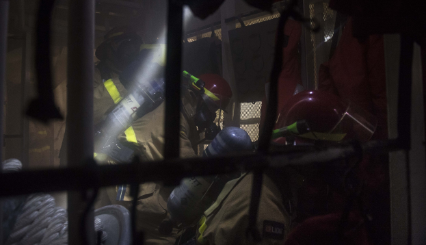 Feuerwehrleute in Schutzausrüstung mit einem, der einen Feuerlöscher hält, einem Metallgrill und einem Seil im Vordergrund und verschiedenen Gegenständen im Hintergrund.