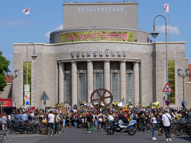 Eine große Gruppe von Menschen versammelte sich vor einem Gebäude mit Säulen und Text, umgeben von Straßenmasten, Lichtern, Schildern, Motorradfahrern, Kraftfahrzeugen, Bäumen und einem bewölkten Himmel während einer Demonstration in Berlin, Deutschland.
