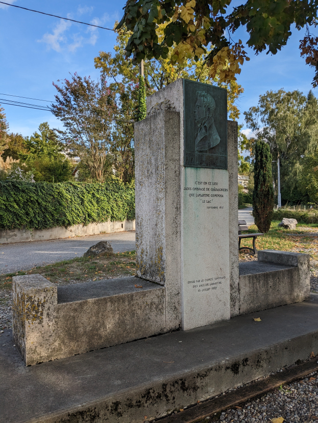 Ein steinernes Denkmal mit eingraviertem Text steht in einem Park, umgeben von Gras, Pflanzen, Bäumen und einer Bank davor, mit einem bewölkten Himmel darüber.
