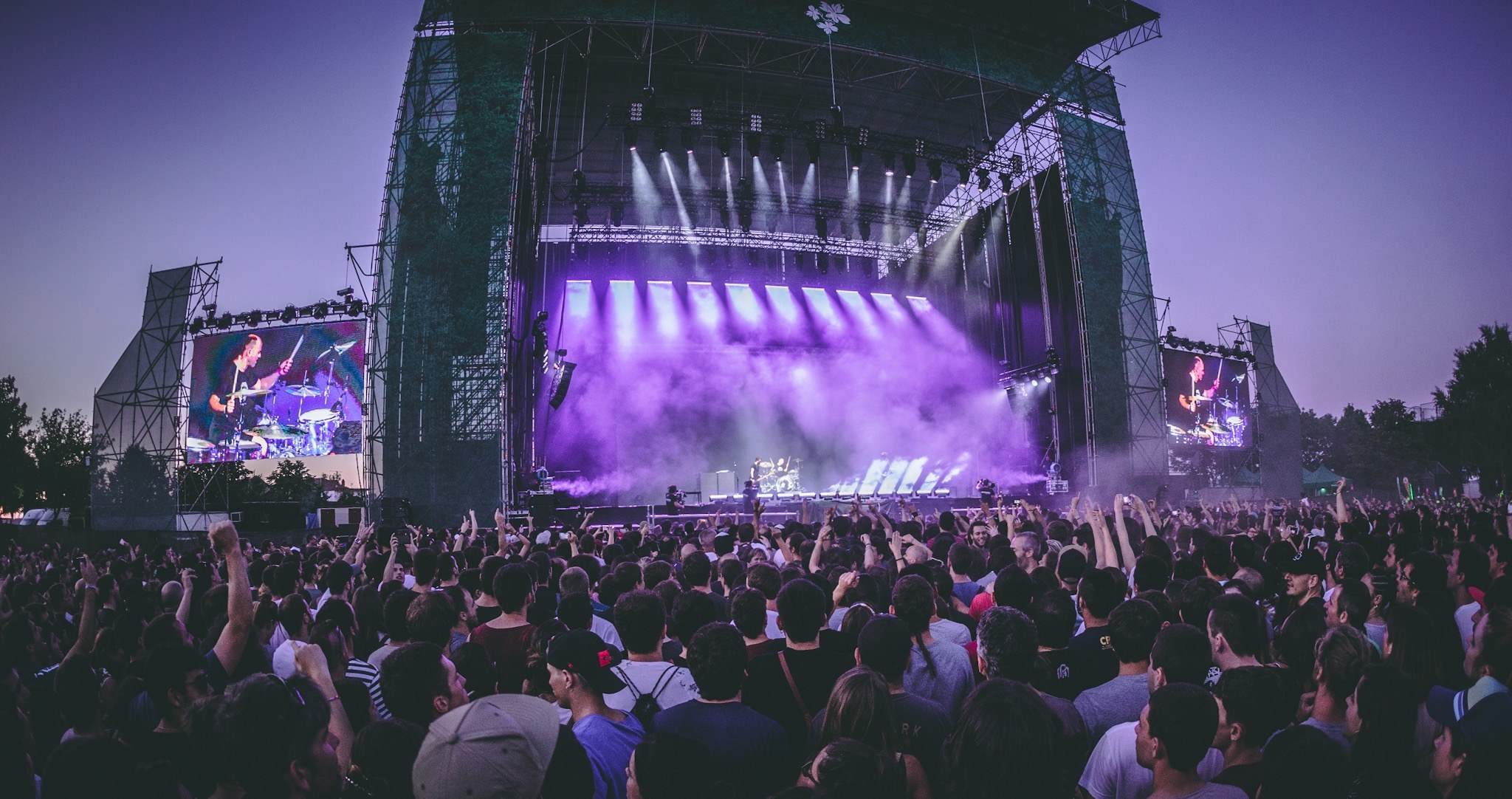 Eine große Menschenmenge bei einem Musikfestival schaut auf der hell erleuchteten Bühne mit Bildschirmen, die vor einem Gebäude, Bäumen und einem klaren blauen Himmel aufgebaut ist.