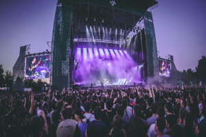 Eine große Menschenmenge bei einem Musikfestival schaut auf der hell erleuchteten Bühne mit Bildschirmen, die vor einem Gebäude, Bäumen und einem klaren blauen Himmel aufgebaut ist.