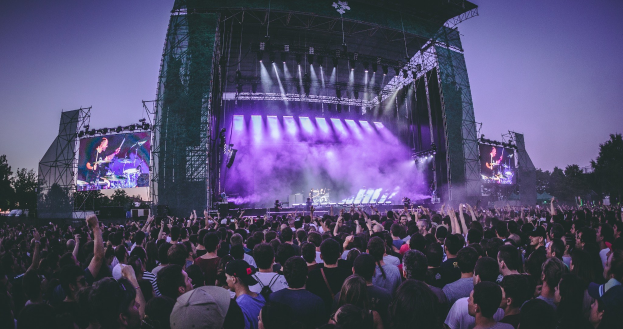 Eine große Menschenmenge bei einem Musikfestival schaut auf der hell erleuchteten Bühne mit Bildschirmen, die vor einem Gebäude, Bäumen und einem klaren blauen Himmel aufgebaut ist.