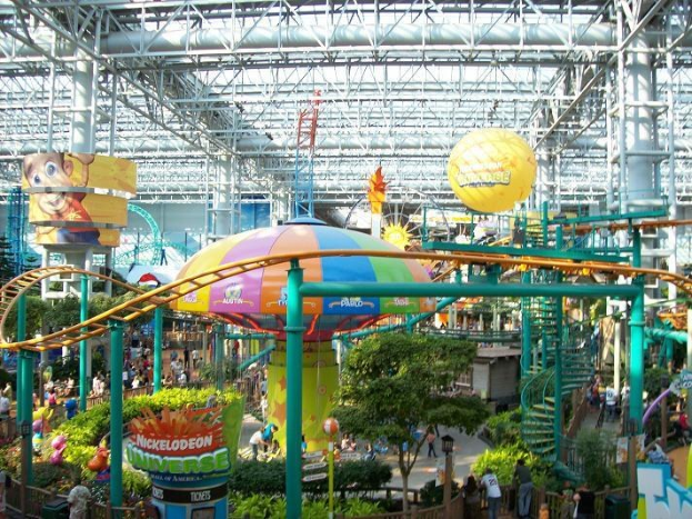 Großer Indoor-Wasserpark mit Achterbahn, umgeben von Menschen, Bäumen, Pflanzen und Beleuchtungskörpern an der Decke.
