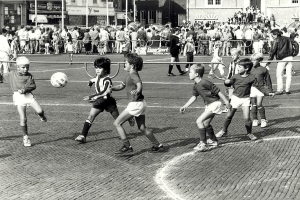 Schwarzes Foto von jungen Jungs, die auf der Straße Fußball spielen, umgeben von einer Zuschauermenge hinter einem Geländer, mit Gebäuden und Polen im Hintergrund.