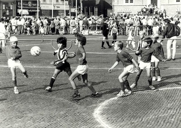 Schwarzes Foto von jungen Jungs, die auf der Straße Fußball spielen, umgeben von einer Zuschauermenge hinter einem Geländer, mit Gebäuden und Polen im Hintergrund.