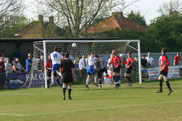 Spieler spielen Fußball auf einem Feld mit einem Tor und Zuschauern hinter ihm, mit Bäumen und Häusern im Hintergrund.