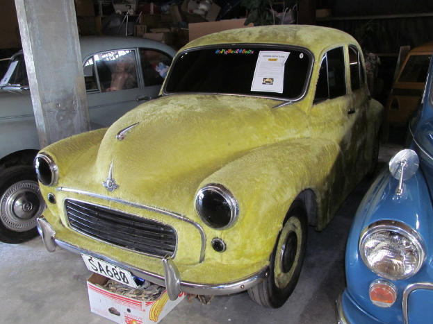 Mehrere Oldtimer sind vor einer Wand mit verschiedenen Zubehörteilen geparkt, links ist eine Säule zu sehen.