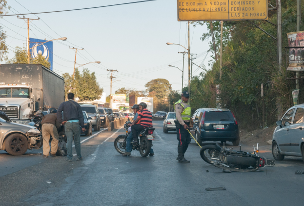 Gruppe von Menschen um ein verunglücktes Motorrad am Straßenrand mit vorbeifahrenden Fahrzeugen, darunter ein Lastwagen, und Hintergrund-elementen wie Bäume, Strommasten und Verkehrszeichen.