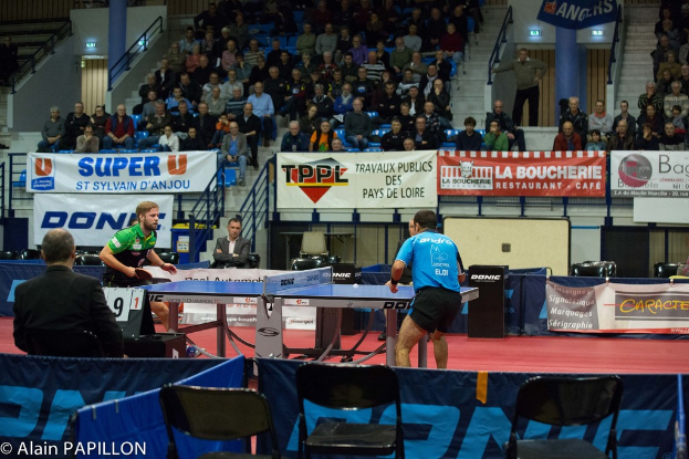Zwei Männer spielen Tischtennis drinnen mit mehreren Zuschauern, die stehen und sitzen, einigen leeren Stühlen und Werbetafeln, mit Text in der unteren linken Ecke.