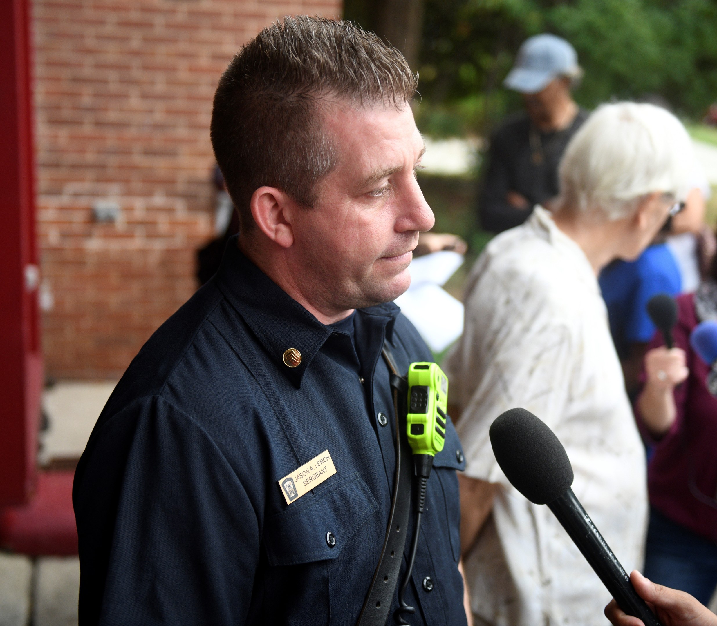 Ein Polizist in Uniform spricht mit Reportern, die ein Mikrofon halten, während andere in der Nähe stehen, vor einer Backsteinwand und Bäumen.