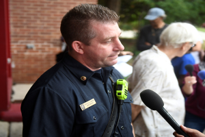Ein Polizist in Uniform spricht mit Reportern, die ein Mikrofon halten, während andere in der Nähe stehen, vor einer Backsteinwand und Bäumen.