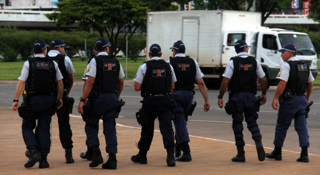Eine Gruppe von Beamten in Uniform, einschließlich Mützen, Hemden, Schutzwesten, Uhren, Hosen und Schuhen, die auf einer Straße mit einem geparkten Lastwagen auf der rechten Seite neben einem Zaun gehen. Im Hintergrund sind Gebäude, Bäume, Pflanzen, Pfosten, Banner und in der oberen rechten Ecke eine Brücke zu sehen.