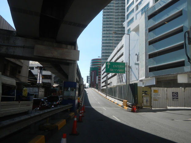 Stadtstraße mit Verkehrskegeln am Straßenrand, fahrende Fahrzeuge, eine Brücke auf der linken Seite, Gebäude mit Namensschildern auf beiden Seiten, Laternenmasten und einen sichtbaren Himmel im Hintergrund.