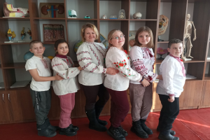 Eine Gruppe von Kindern in traditioneller ukrainischer Kleidung vor einem Bücherregal mit Spielzeug, einem Skelett und anderen Gegenständen in einer scheinbar ukrainischen Schulumgebung.