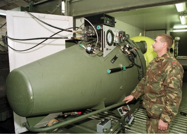 Ein Mann in militärischer Uniform steht neben einer großen grünen Bombe in einer unordentlichen industriellen Umgebung mit Maschinen, Drähten und beleuchteten Wänden.