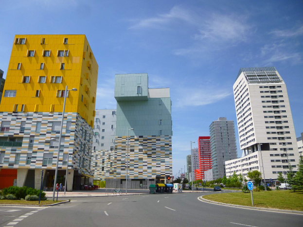 Eine Stadtstraße mit hohen, einzigartig gestalteten Gebäuden der Universität Rotterdam im Hintergrund, Straßenlaternen, Straßenschildern, Kraftfahrzeugen, Infotafeln, Büschen, Bäumen und einem bewölkten Himmel.