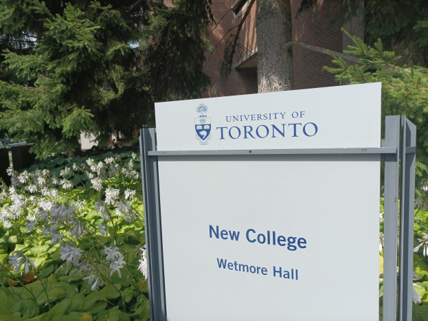Außenansicht des neuen Colleges der Universität Toronto an Wetmore Hall, mit einem Schild vor dem Gebäude umgeben von grünem Gras, bunten Blumen, hohen Bäumen und einem hellblauen Himmel.