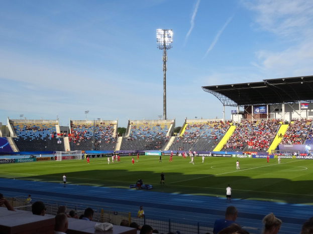 Ein Fuballspiel in einem gro├čen, beleuchteten Stadion mit Zuschauern auf den R├Ąngen und Spielern auf dem Feld unter einem sichtbaren Himmel.