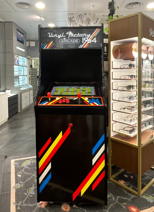 Ein Geschäft mit einem vintage Arcade-Automaten in der Mitte, einem Glastisch mit einer Vitrine auf der rechten Seite und einer retro-stylischen Beleuchtung mit Schränken und einem Fernseher im Hintergrund.