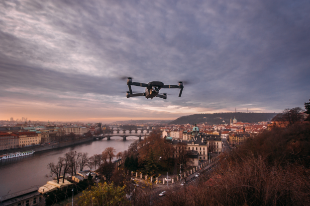 Eine Drohne fliegt über einer Stadt mit Gebäuden, Bäumen und Fahrzeugen, wobei ein Fluss und eine Brücke im Hintergrund zu sehen sind.