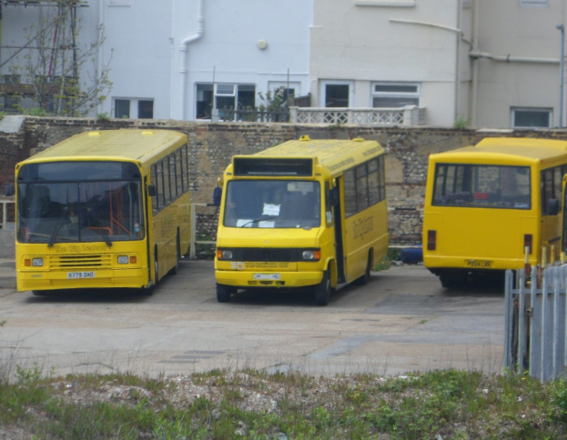 Drei gelbe Busse nebeneinander auf einem eingezäunten Parkplatz mit Gras, einem Gebäude und anderen Strukturen geparkt