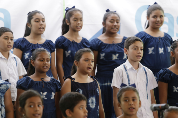 Gruppe von Kindern singt vor einem Mikrofon, mit einem Schulchor-Banner im Hintergrund.