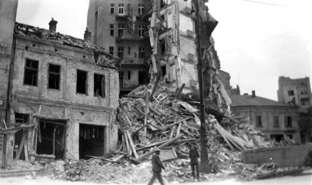 Ein Schwarz-Weiß-Foto eines Mannes, der durch die Trümmer eines zerstörten Gebäudes geht, umgeben von anderen Gebäuden mit Fenstern, Pfählen und anderen Gegenständen, mit dem Himmel im Hintergrund.