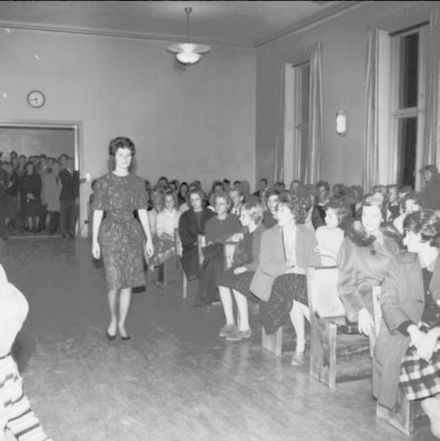 Eine Frau in einem schwarzen und weißen Kleid läuft über den Laufsteg während einer Modenschau für Frauenrechte, mit einer Menge, die sitzt und zusieht, einer Uhr und Lampen an der Wand und einer beleuchteten Decke.