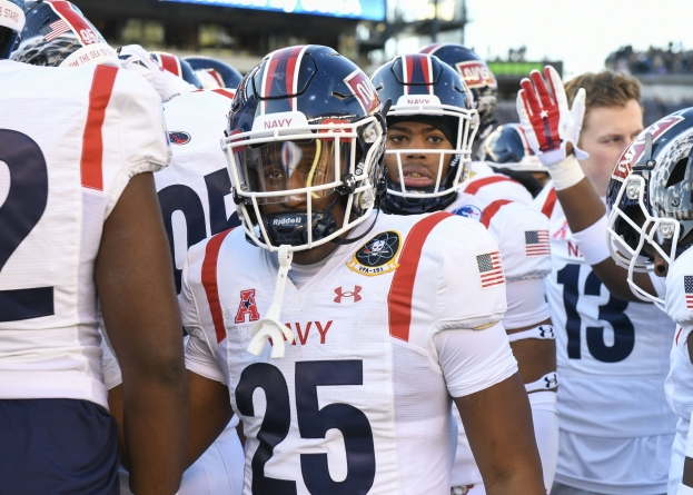 Gruppe von Männern in Football-Uniformen auf einem Feld mit Helmen und Handschuhen, mit einer unscharfen Menge, einem Anzeigebrett mit 'Auburn vs Virginia Tech - 11/28/15 College Football Pick, Odds, and Prediction' und Himmel im Hintergrund.