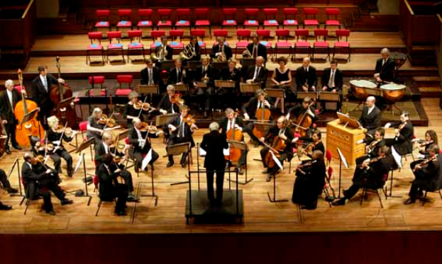 Ein großes Orchester auf einer Bühne mit einem Dirigenten vorne, der verschiedene Instrumente spielt, mit Ständern und anderen Gegenständen drumherum und Stühlen und Treppen im Hintergrund.