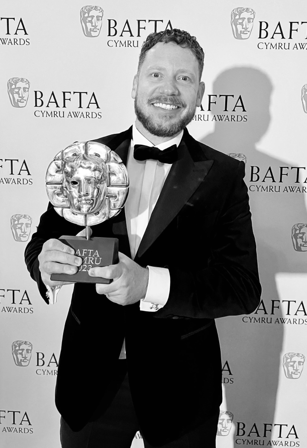 Ein Mann in einem Smoking, der eine BAFTA-Auszeichnung hält und vor einer Tafel mit der Aufschrift "BAFTA Awards 2018" steht.