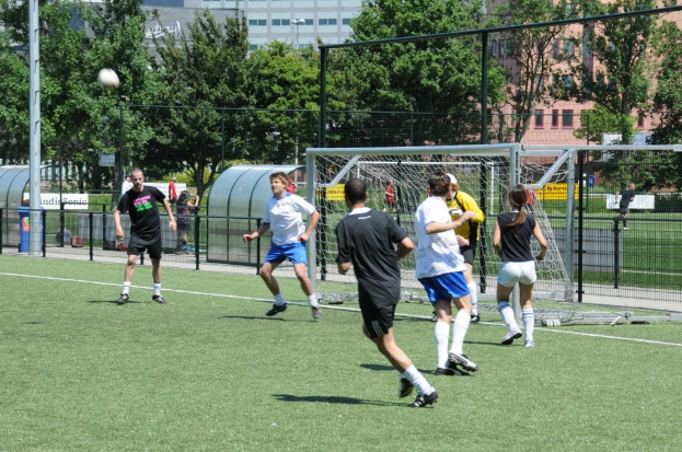 Menschen in Kleidung, die Fußball spielen, auf einem Feld mit einem Zaun und einem Tor in der Mitte, einem Pfosten und einem Fußball oben links und Bäumen und Gebäuden im Hintergrund.