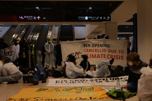 Eine Gruppe von Menschen sitzt auf dem Boden eines Flughafens und hält ein Banner mit der Aufschrift "Berlin wegen Klimakrise gestrichen". Um sie herum liegen Taschen, Flaschen und andere Gegenstände verstreut, im Hintergrund sind Rolltreppen, Tafeln mit Text und Deckenleuchten zu sehen.