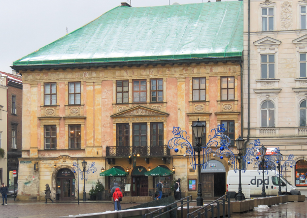 Großes Gebäude mit grüner Dachfläche in einer Stadt, umgeben von Straßenmasten, Laternen, Fahrzeugen, Fußgängern, Geländern, Bäumen und einem klaren Himmel.