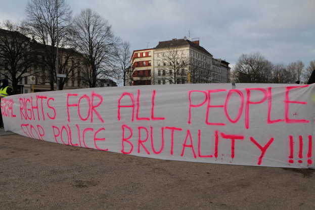 Eine Gruppe von Menschen steht auf dem Boden und hält ein Banner mit der Aufschrift "Rechte für alle Menschen Stoppt Polizeigewalt", mit einem Straßenschild, einem Schild, Bäumen, Gebäuden mit Fenstern und einem bewölkten Himmel im Hintergrund.
