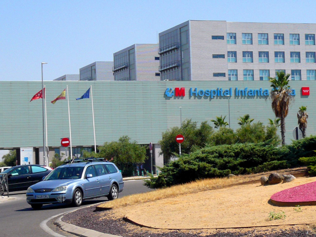 Krankenhaus mit geparkten Autos, umgeben von städtischer Infrastruktur und Grün unter einem klaren blauen Himmel.