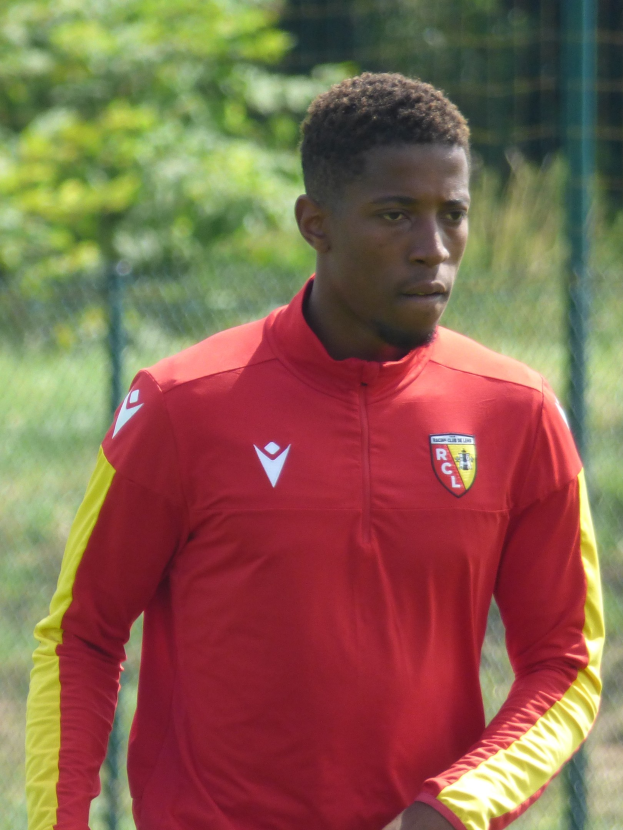 Ein junger Mann in einem roten und gelben Fußballtrikot steht auf einem Feld und hält den Zaun mit konzentrierter Miene fest.