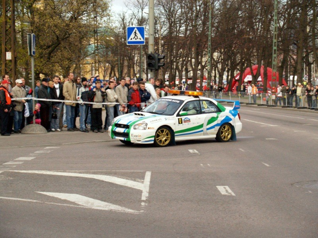 Ein Rallyefahrzeug fährt an einer von Zuschauern gesäumten Straße vorbei, an der Bäume, Pfosten, Verkehrszeichen, Geländer, Gebäude und ein klarer blauer Himmel zu sehen sind.