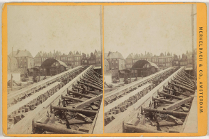 Ein altes Schwarz-Weiß-Foto einer Bahnschiene mit Gebäuden im Hintergrund, Text auf der rechten Seite lesbar "Merkelbach & Co, Co. Amsterdam" und verstreute hölzerne Gegenstände im Vordergrund.