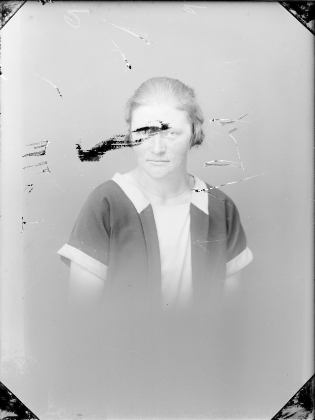 Schwarz-weißes Foto einer Frau mit einem zerbrochenen Glasfenster im Hintergrund, die direkt in die Kamera schaut und mit leicht geöffneten Lippen, zurückgekämmtem Haar und gefalteten Händen dasteht.